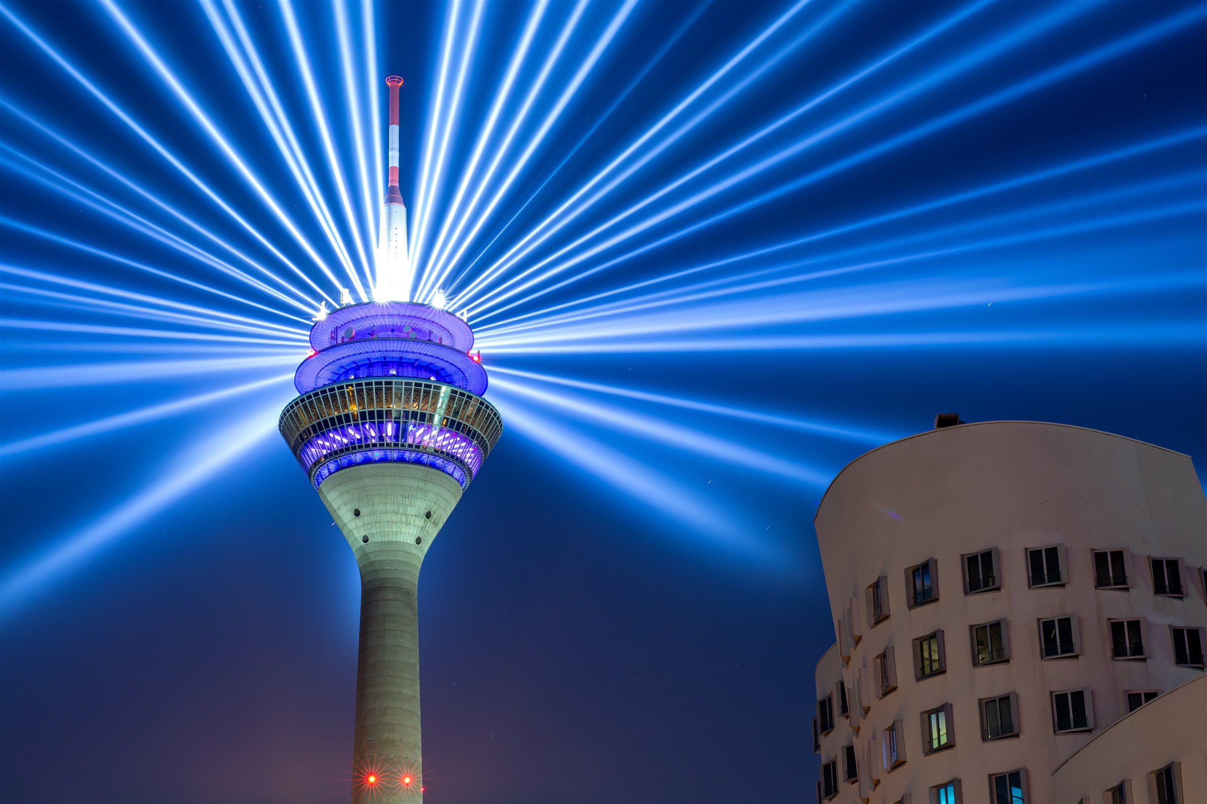 Blaue Lichtstrahlen fächern sich über dem Rheinturm auf, daneben ein geschwungenes Gebäude