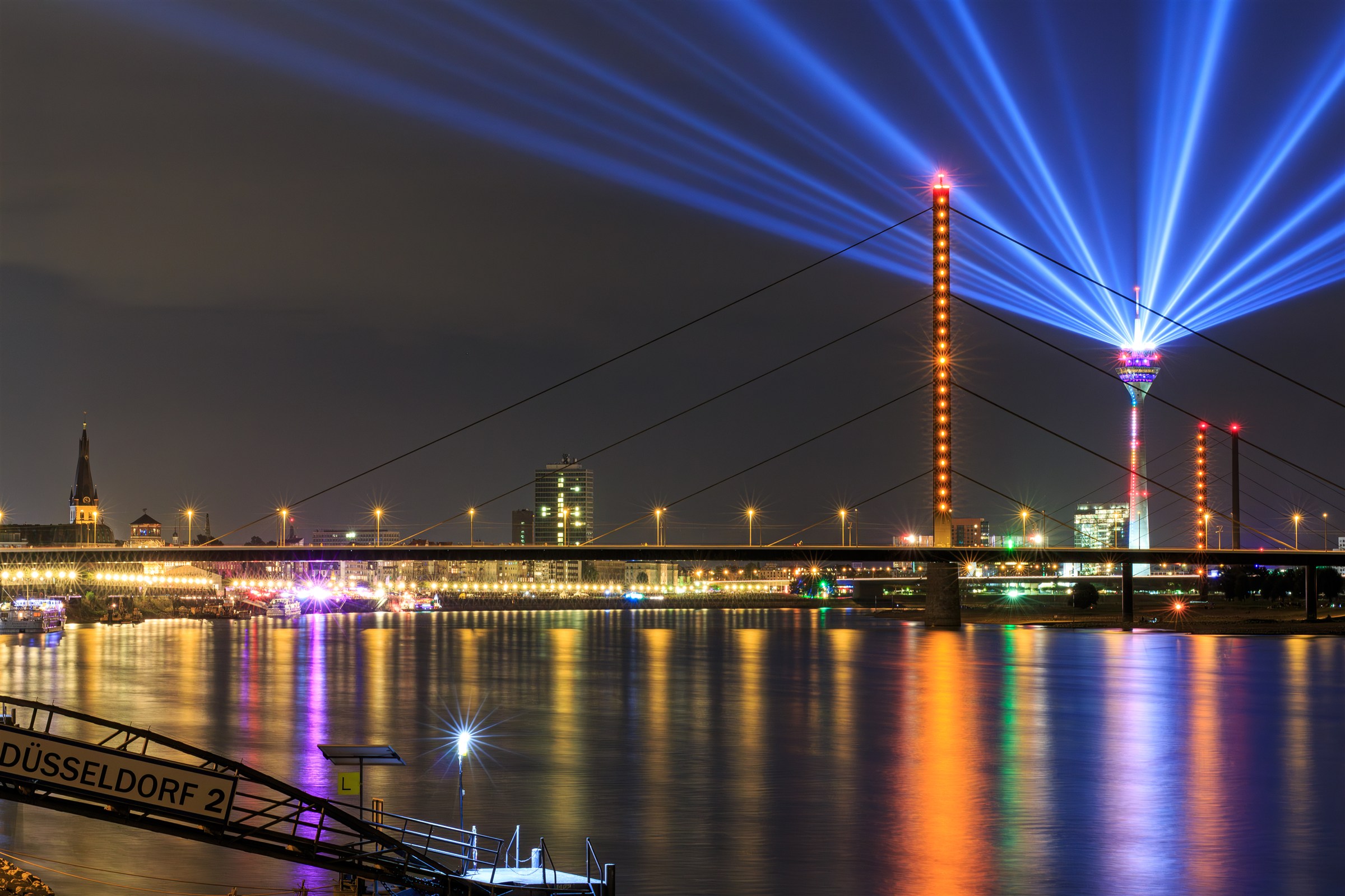 Nächtliche Rheinansicht mit beleuchteter Brücke, Lichtstrahlen am Rheinturm und Spiegelungen im Wasser